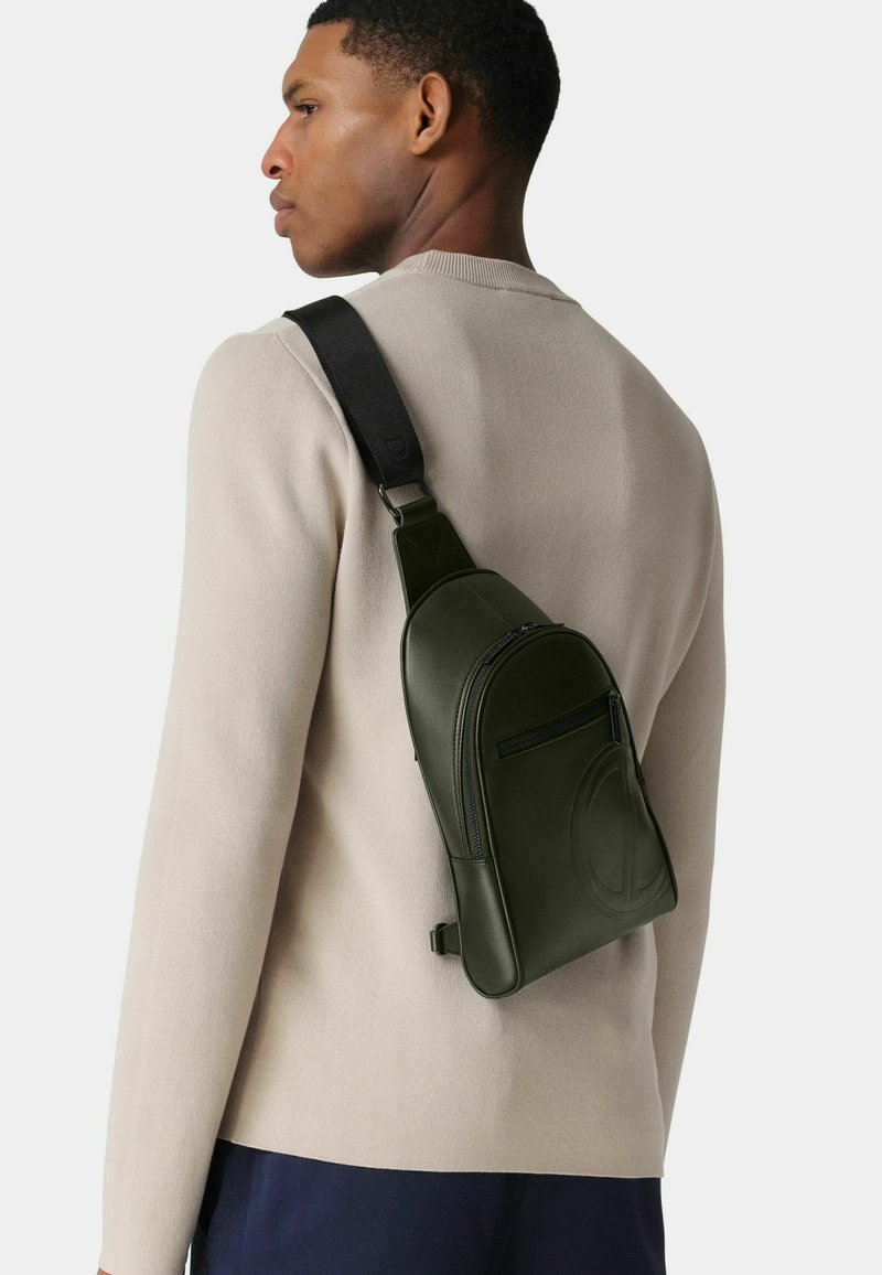 Grüne Leder-Umhängetasche mit einer vorderen Reißverschlusstasche und geprägtlichem Logo, getragen über einem beigen Pullover. Verstellbarer schwarzer Riemen für Komfort.