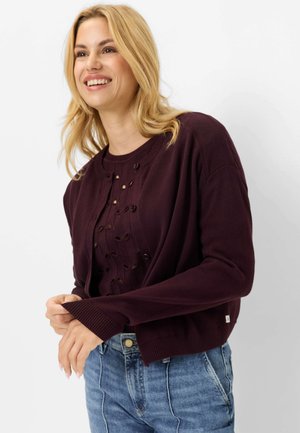 Smilende blond kvinde iført en mørkelilla knap-cardigan og en matchende top med mønstrede huller, kombineret med blå denimjeans.