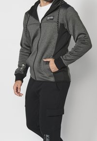 Veste zippée pour homme en gris foncé et noir, fabriquée dans un tissu mélangé, avec capuche et détails logo sur la poitrine et la manche. Associée à un pantalon noir.