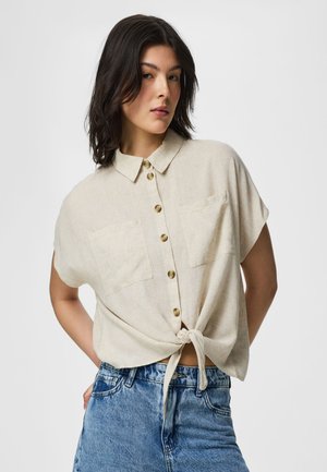 Tummatukkainen nainen, jolla on beige lyhythihainen napillinen paita sidottuna vyötärölle ja siniset farkut, seisoo kädet selän takana.