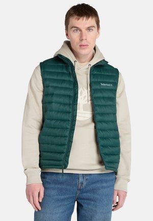 Grøn quiltet pufvest med horisontal quiltet tekstur, front lynlås og Timberland-logo. Bæres over en cremefarvet hættetrøje og parret med blå jeans.
