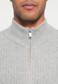 Camisola cinzenta canelada com gola alta e fecho prateado. Tecido texturizado com padrões verticais que se estendem até ao pescoço.