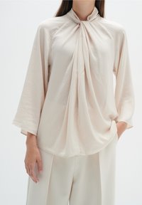 Blusa beige chiaro con un davanti drappeggiato e colletto alto. Maniche lunghe e larghe, realizzate in un tessuto morbido e fluido, abbinate a pantaloni a vita alta.