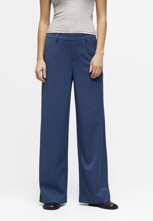 Vrouw met blauwe broek met wijde pijpen, grijze mouwloze top en zwarte schoenen, staand tegen een effen witte achtergrond.