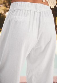Pantaloni in lino bianco con vita elastica arricciata e cintura decorativa. Presentano due tasche posteriori e un tessuto leggero e strutturato.