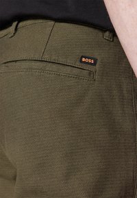 Beżowe spodnie z subtelnym wzorem w jodełkę, z tylną kieszenią i małą czarną metką z pomarańczowym logo „BOSS”.