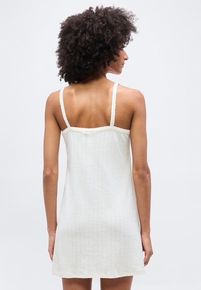 Γυναίκα με σγουρά μαλλιά που φοράει ένα αμάνικο, λευκό, ριμπ φόρεμα, απεικονιζόμενη από πίσω σε απλό ανοιχτό φόντο.