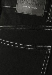 Närbild av svart denimtyg med vit sömnad och en svart läderlapp märkt "bershka DENIM COLLECTION."