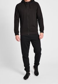Svart zip-up huvtröja med snörning i huvan, tillsammans med matchande svarta joggers. Båda plagg är gjorda av mjukt, texturerat tyg. Svarta sportskor fulländar looken.
