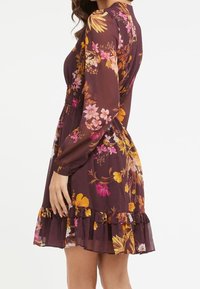 Robe florale en bordeaux foncé avec des motifs floraux roses et jaunes. Manches longues, taille cintrée et détails de bas à volants superposés.
