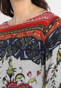 Pull gris avec des motifs floraux et géométriques complexes en rouge, bleu, vert et jaune, avec un décolleté large et une texture douce.