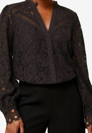 Blouse en dentelle florale noire avec boutons sur le devant, col en V, manches longues bouffantes et poignets en dentelle détaillée, portée avec un pantalon noir.