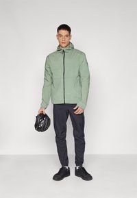 Lys grøn, letvægts zip-up jakke med hætte, parret med mørke bukser og sorte sko. Modellen holder en sort cykelhjelm.