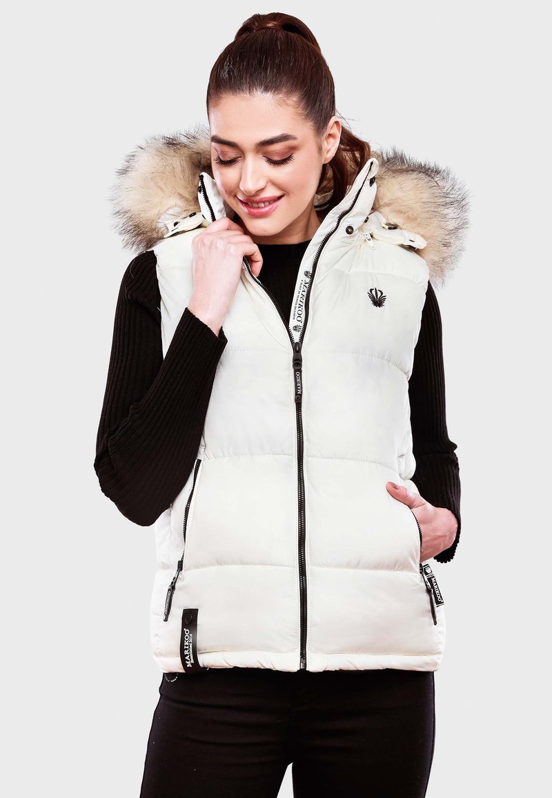 Gilet in piuma bianco con cappuccio rimovibile, dotato di bordatura in pelliccia sintetica, due tasche laterali e zip frontale. Maglione nero a costine indossato sotto.