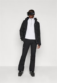 Casaco isolado preto com fecho, top térmico branco, calças de esqui pretas e botas de esqui pretas. Modelo a usar óculos de esqui pretos. Design simples.