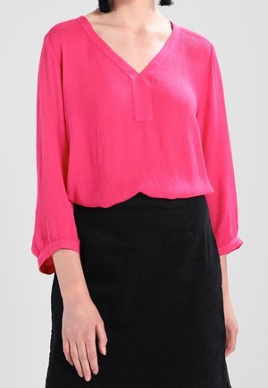 Femme portant un blouse V à manches longues rose vif et une jupe noire simple sur un fond neutre.