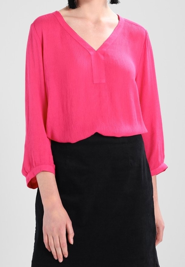 Donna che indossa una blusa a maniche lunghe col collo a V di un brillante rosa e una gonna nera semplice, su uno sfondo neutro.