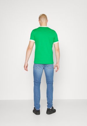 Mężczyzna o krótkich blond włosach, ubrany w zielony t-shirt, niebieskie dżinsy i czarne tenisówki, stoi tyłem na jednolitym białym tle.