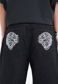 Svarta denimjeans med silverbrodering av ett tigerhuvud på båda bakfickorna. Tyget är robust och har en klassisk femfickdesign.
