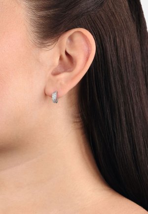 Primo piano di un orecchio sinistro con capelli scuri e lisci, che indossa un piccolo orecchino a cerchio d'argento decorato con gemme trasparenti.