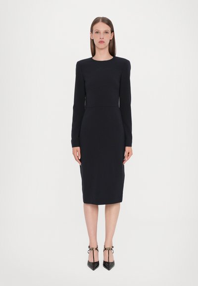 Robe noire ajustée à manches longues avec un col rond. Texture lisse, longueur aux genoux et détails de couture subtils à la taille.