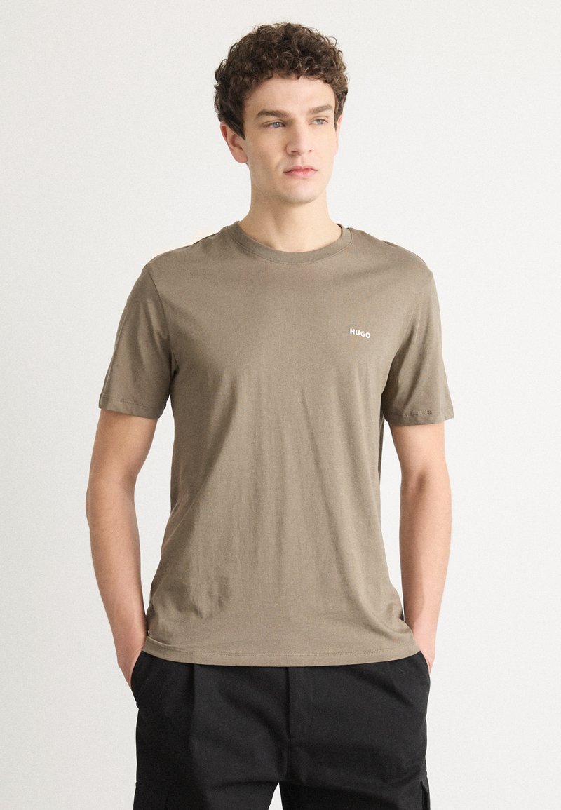 T-shirt a maniche corte, con scollo rotondo, in tessuto taupe con una texture liscia. Presenta un piccolo logo bianco "HUGO" sul petto.