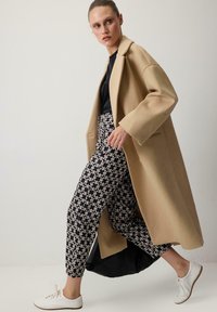 Beige wool coat with a relaxed fit, paired with black and white patterned trousers. White sneakers complete the outfit.