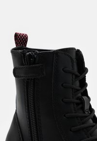 Bottine en cuir noir avec lacets, fermeture éclair sur le côté et une languette en relief rouge et noire. Fini lisse et couture détaillée.