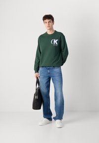 Hanorac verde cu guler rotund, cu logo alb CK, blugi denim albaștri și adidași albi; ținând o geantă neagră cu două fermoare superioare.