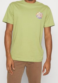 Světle zelené bavlněné tričko s krátkými rukávy, kulatým výstřihem a kruhovou vícebarevnou grafikou „Santa Cruz“ na levé straně hrudníku.