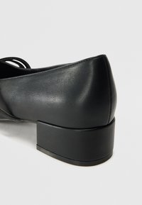 Scarpe in ecopelle nera con tacco inclinato, texture liscia e dettagli con lacci sui lati per un tocco di design in più.