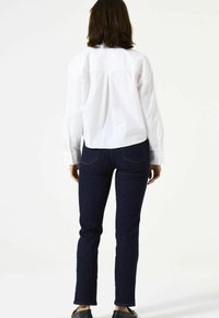 Chemise blanche à col, manches longues, détail de la pièce d'épaule arrière ; associée à un jean slim bleu foncé. Design minimaliste, sans motifs ni éléments métalliques visibles.