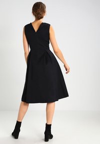 Robe noire en A avec décolleté en V et tissu texturé, taille cintrée et plissée. Fermeture éclair au dos et longueur au-dessus du genou. Associée à des bottines.