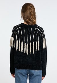 Schwarzer Pullover mit strukturiertem Strick, der cremefarbene Fransen über den Rücken hat, sowie gerippte Bündchen und Saum. Kombiniert mit blauen Jeans.