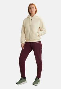 Felpa con zip color crema, dotata di cappuccio e tasche anteriori, abbinata a joggers a gamba affusolata color bordeaux e scarpe da ginnastica verdi.