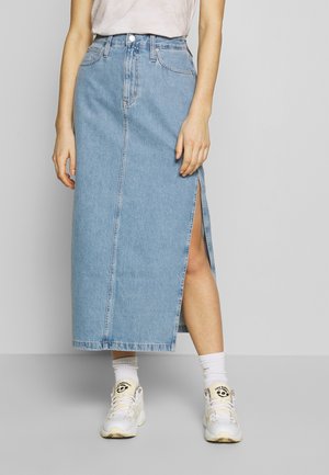 Vaalea sininen denim-maxihame korkealla halkiolla, jossa on viisi taskua, niittiyksityiskohtia ja suora leikkaus. Yhdistettynä valkoisiin tennareihin.