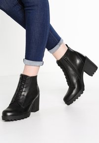 Botas de cuero negras hasta el tobillo con un alto tacón cuadrado, parte frontal con cordones, cremallera lateral y suela robusta. Puños de mezclilla enrollados visibles.