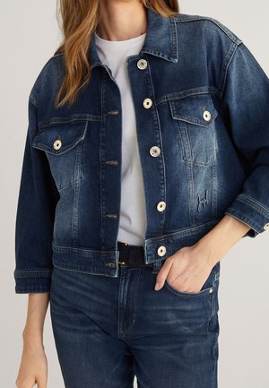 Femme portant une veste en jean bleu foncé avec des poches à rabat boutonnées sur une chemise blanche et un jean bleu foncé assorti, main dans la poche.
