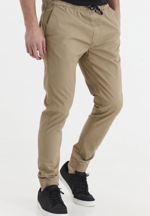 Pantalones jogger beige hechos de tela con una cintura elástica, cordón ajustable y tobillos ceñidos, combinados con zapatillas negras.