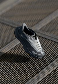 Shoe sportiv gri și negru cu fermoar frontal, partea superioară texturată și talpă Vibram, având un design aerodinamic și o formă modernă.