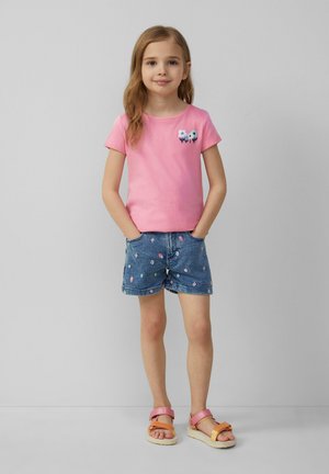 Jong meisje met lang haar, gekleed in een roze bloemen t-shirt, blauwe denim shorts met kleine prints en meerkleurige sandalen, staand met handen in de zakken.
