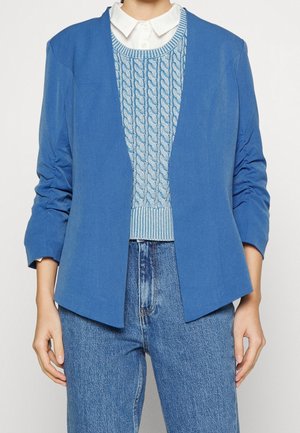 Personne portant une chemise blanche avec col, un pull bleu en maille torsadée, un blazer bleu et un jean bleu taille haute, les mains détendues le long du corps.