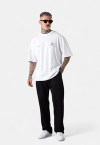 Mann mit Tattoos und Sonnenbrille, der ein weißes, übergroßes T-Shirt, schwarze Hose und weiße Sneaker trägt und mit einer Hand in der Tasche steht.