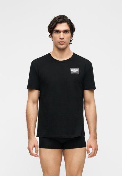 Άνδρας με σκούρα μαλλιά φοράει μαύρο t-shirt Moschino Active και μαύρα μποξεράκια σε απλό λευκό φόντο.