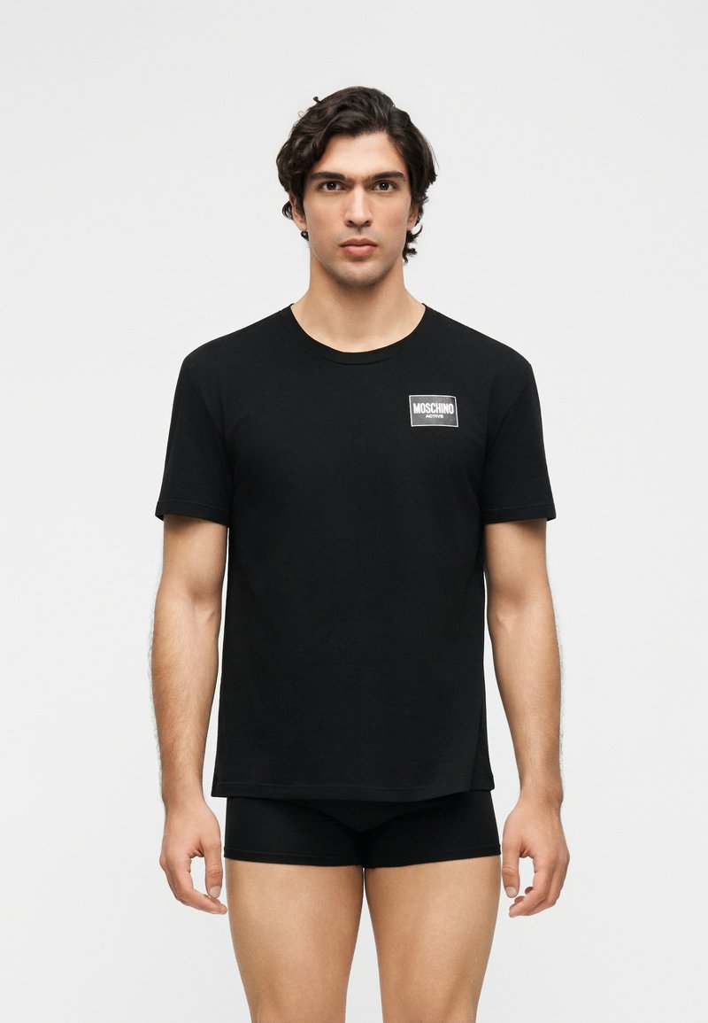 Άνδρας με σκούρα μαλλιά φοράει μαύρο t-shirt Moschino Active και μαύρα μποξεράκια σε απλό λευκό φόντο.