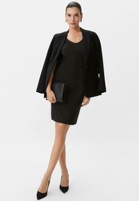 Schwarzes, figurbetontes Kleid mit V-Ausschnitt, kombiniert mit einem taillierten schwarzen Blazer. Das Model hält eine schwarze Clutch und trägt spitze Pumps.