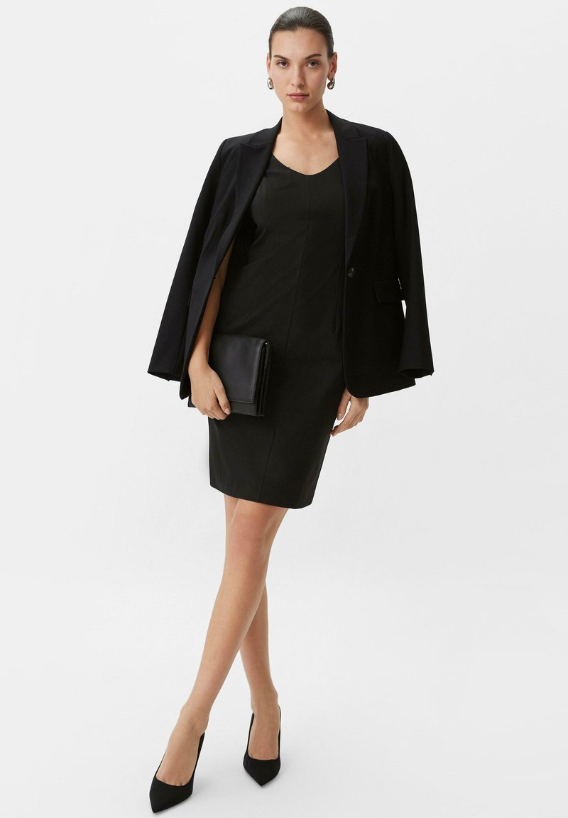Schwarzes, figurbetontes Kleid mit V-Ausschnitt, kombiniert mit einem taillierten schwarzen Blazer. Das Model hält eine schwarze Clutch und trägt spitze Pumps.