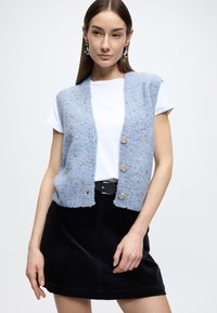 Lichtblauw gebreid vest met een gestructureerde afwerking en spikkels, vijf knopen, gedragen over een wit T-shirt en gecombineerd met een zwarte corduroy rok.