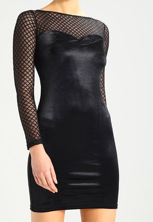 Femme portant une mini-robe en velours noir avec des manches longues transparentes présentant un motif en maille losange.