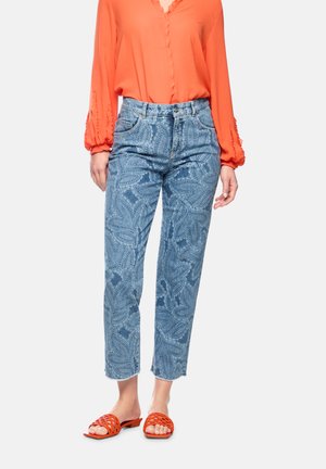 Woman wearing blue patterned jeans, an orange long-sleeve blouse, and orange open-toe woven sandals standing on a white background.